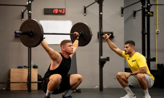 Athlete performing weightlifting exercise under trainer's guidance in a gym setting.